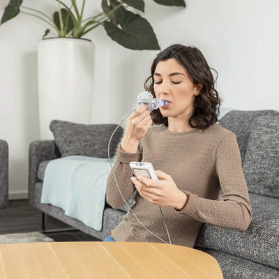 Person sitzt auf einem Sofa und inhaliert mit einem eFlow Vernebler, während sie in der Anderen Hand die Steuereinheit hält